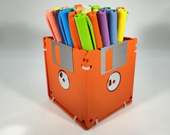 May include: A square pen holder constructed from orange floppy disks joined with white plastic ties. The container is filled with a variety of colorful felt-tip markers, including shades of lime green, bright orange, purple, blue, and yellow.