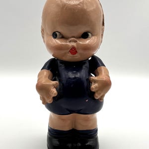 May include: Vintage ceramic doll with a bald head, dark eyes, and red lips. The doll is wearing a dark blue outfit and black shoes. The doll has a stern expression and is standing upright.