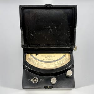 Puede incluir: Medidor de amperios de CA General Electric negro en una caja con bisagras. La esfera del medidor es de color crema con marcas negras y un indicador de aguja. El medidor lee hasta 1500 A.C. Amperios.