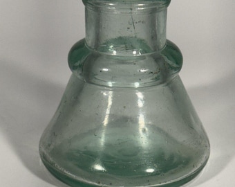 May include: A clear, light green glass inkwell with a wide, conical base that tapers upward to a cylindrical neck. The glass features a smooth, rounded shoulder and a thick, reinforced rim at the top opening, showing subtle surface imperfections.