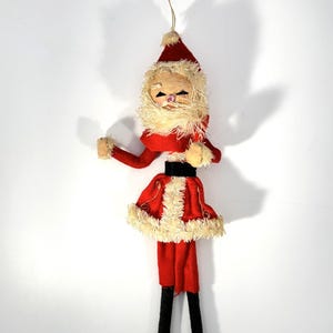 May include: A vintage Santa Claus Christmas ornament. The ornament features a red felt suit with a black belt, a white beard, and a red hat. The arms and legs are made of felt and the face is made of a light-colored material.