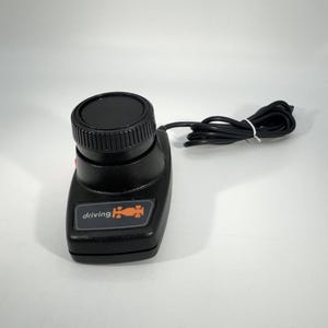 May include: Black driving controller with a round top and a cord. The controller has the word "driving" and a race car graphic in orange. The controller is on a white background.