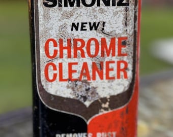 May include: A vintage rectangular metal Simoniz Chrome Cleaner can with a black and red color scheme. The front features the text SIMONIZ, NEW!, CHROME CLEANER, and REMOVES RUST CLEANS POLISHES. The surface shows significant wear and rust.