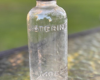 May include: A clear glass bottle with a black screw-on cap. The bottle features embossed text reading LISTERINE on the upper body and LAMBERT PHARMACAL COMPANY on the lower body. The container has a cylindrical shape with a narrow neck.