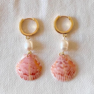 Op de afbeelding: Een paar gouden creolen met een parel en een roze schelpbedel die aan elke creool hangt.