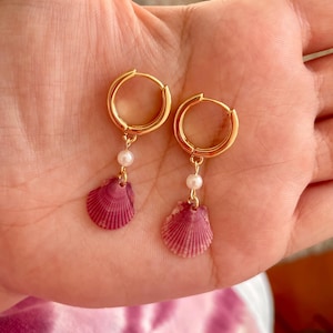 May include: A pair of gold hoop earrings featuring a small white pearl bead and a dangling pink scallop shell charm. The earrings have a hinged closure mechanism and are designed with a polished metallic finish on the circular hoops.