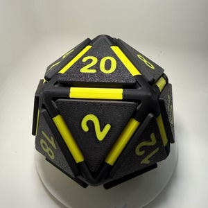 May include: A large, black and yellow twenty-sided die with yellow numbers. The die has a textured surface and is designed for tabletop gaming. The numbers are in a bold, easy-to-read font.
