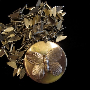 Op de afbeelding: Een gouden ketting met een medaillon met een vlinderbedel. De ketting is gemaakt van een ketting met kleine vlinderbedels. Het medaillon is rond en heeft een vlinderontwerp aan de voorkant.