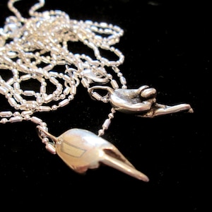 May include: A silver chain necklace with two pendants. One pendant is a small, abstract shape, and the other is a hand with the fingers curled into a fist.