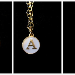 May include: Three gold necklaces with a round pendant featuring the letter A. The pendants are black, white, and pink.