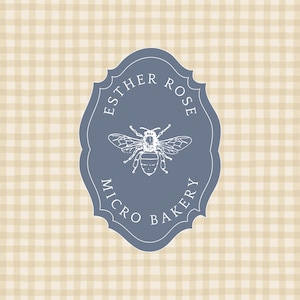 Puede incluir: Una etiqueta azul con el texto "Esther Rose Micro Bakery" y una ilustración detallada de una abeja. La etiqueta tiene un borde decorativo y está sobre un fondo a cuadros beige y blanco.