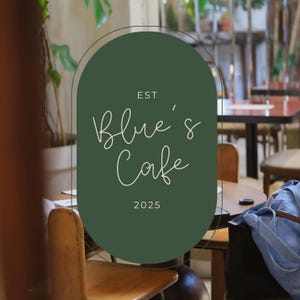 Puede incluir: Un letrero ovalado de color verde oscuro con el texto en blanco EST Blue's Cafe 2025 en una fuente manuscrita fina y elegante. El letrero está enmarcado por un borde negro fino y está colocado en el centro, mostrando el nombre y el año.