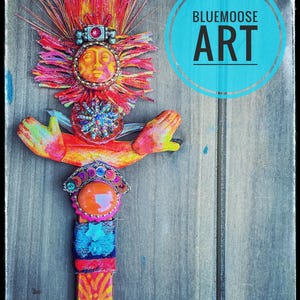 May include: A mixed-media wall hanging featuring a central sun face with orange and red fringe, outstretched hands, a large orange cabochon, and colorful fabric accents. The piece includes a blue circular logo with the text BLUEMOOSE ART.