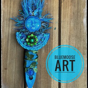 May include: A blue decorative wall hanging featuring a central sun face with a butterfly accent, surrounded by blue fringe. The piece includes a green beaded medallion, green leaf embellishments, and a blue circular stone, with BLUEMOOSE ART text.