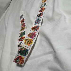 May include: White ribbon with colorful cartoon character designs and a clear plastic clasp. The ribbon features various cartoon characters in a repeating pattern. The ribbon is laying on a white surface.