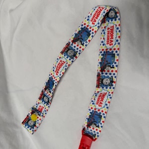 May include: A Thomas & Friends themed dummy clip. The clip features a repeating pattern of the blue train character, Thomas, on a white background with red and blue polka dots. The clip has a red plastic clasp and a yellow button.