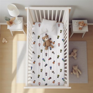 Könnte beinhalten: Ein weißes Babybett mit einem gemusterten Spannbettlaken mit bunten Zeichentrickfiguren. Ein brauner Teddybär und ein weißes Kissen liegen auf dem Laken. Das Bettchen ist von kleinen weißen Tischen und einem grauen Teppich umgeben, mit zusätzlichen Teddybären.