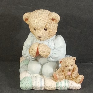 May include: A ceramic figurine of two teddy bears. The larger bear is kneeling on a patchwork quilt and has its paws clasped in prayer. The smaller bear is sitting next to the larger bear and is holding a heart in its paws.