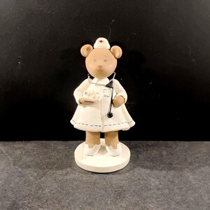May include: A figurine of a bear dressed as a nurse, wearing a white coat with blue stitching and a nurse's cap. The bear holds a tray with small objects and a stethoscope. The figurine stands on a white circular base.