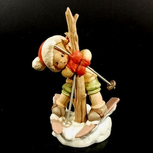 May include: A ceramic figurine of a child skiing, wearing a red hat and scarf, green pants, and tan boots. The figure is posed on a snowy base with skis and poles, leaning against a wooden tree trunk. The figurine is a winter-themed collectible.