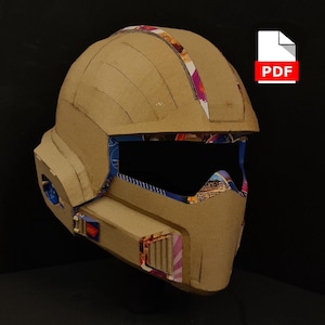 以下が含まれることがあります： 未来的なデザインの手作り段ボールヘルメット。ヘルメットは主にタン色で、バイザーと上部にカラフルな模様のアクセントが付いています。PDFアイコンはデジタルテンプレートを示唆しています。