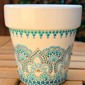 May include: A cream-colored terracotta plant pot featuring a hand-painted mandala design in shades of teal, turquoise, and metallic gold. The pattern consists of intricate dot work, scalloped arches, and small embedded crystals.