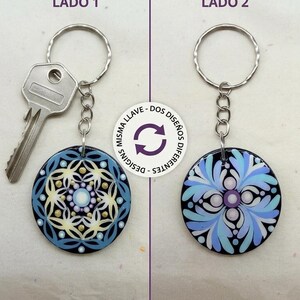 Puede incluir: Un llavero circular de mandala de doble cara con dos diseños distintos. El lado 1 muestra un mandala geométrico en azul, amarillo y blanco. El lado 2 presenta un diseño floral en azul y morado. Incluye una anilla metálica y una llave plateada.
