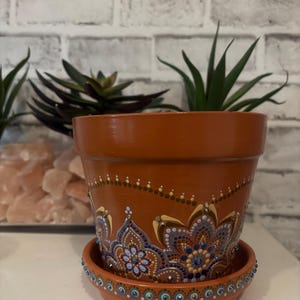 May include: Terracotta-colored flower pot with a matching saucer. The pot is adorned with a hand-painted mandala design in shades of blue, gold, and white. The saucer's rim is decorated with small, blue-colored jewels. The pot is filled with a green plant.