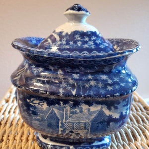 May include: A blue and white porcelain sugar bowl with a lid. The bowl has a floral pattern and a scene of a house with trees. The bowl is sitting on a wicker surface.