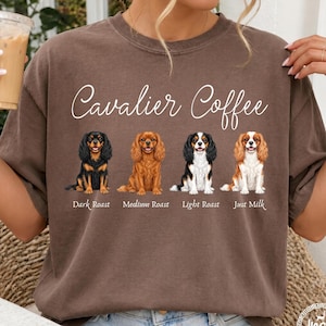 Peut inclure: Un t-shirt marron à manches courtes avec le texte "Cavalier Coffee" au-dessus de quatre illustrations de chiens Cavalier King Charles. Chaque chien est accompagné du texte "Dark Roast", "Medium Roast", "Light Roast" et "Just Milk".