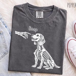 Peut inclure: Un t-shirt gris anthracite avec un motif blanc représentant une main squelettique touchant le nez d'un chien squelettique. Le texte Boop est imprimé au-dessus du chien. Le t-shirt porte une étiquette Comfort Colors au col et est plié.