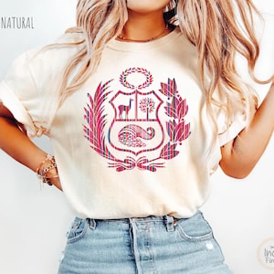 Peut inclure: T-shirt crème avec un motif d'armoiries péruviennes en tartan rouge et bleu. Les armoiries comprennent un lama, un arbre et une corne d'abondance. Le mot "NATURAL" est imprimé en haut.