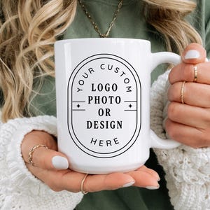 Puede incluir: Una taza de cerámica blanca con un marco ovalado impreso en negro con el texto YOUR CUSTOM LOGO PHOTO OR DESIGN HERE. La taza tiene un asa grande en el lado derecho y está sostenida por dos manos, mostrando un diseño limpio para personalizar.