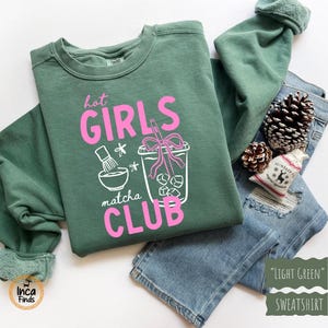 May include: Light green sweatshirt with the text "Hot Girls Matcha Club" in pink. The graphic includes a matcha bowl, a drink with ice, and a straw. The sweatshirt is folded on top of blue jeans, with pine cones and a small gift bag.