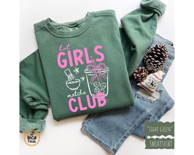 May include: Light green sweatshirt with the text "Hot Girls Matcha Club" in pink. The graphic includes a matcha bowl, a drink with ice, and a straw. The sweatshirt is folded on top of blue jeans, with pine cones and a small gift bag.