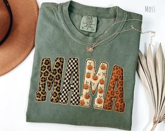 Pode incluir: Camiseta verde musgo com a palavra "MAMA" em um design de patchwork. As letras apresentam estampas de leopardo, xadrez preto e branco e padrões de abóbora. Um colar de prata com um pingente repousa sobre a camisa.