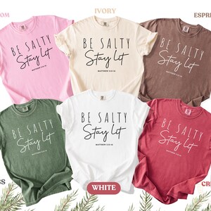 May include: A collection of six short-sleeved t-shirts in various colors: blossom pink, ivory, espresso brown, moss green, white, and crimson red. Each shirt features the text "BE SALTY Stay Lit" in a handwritten style.