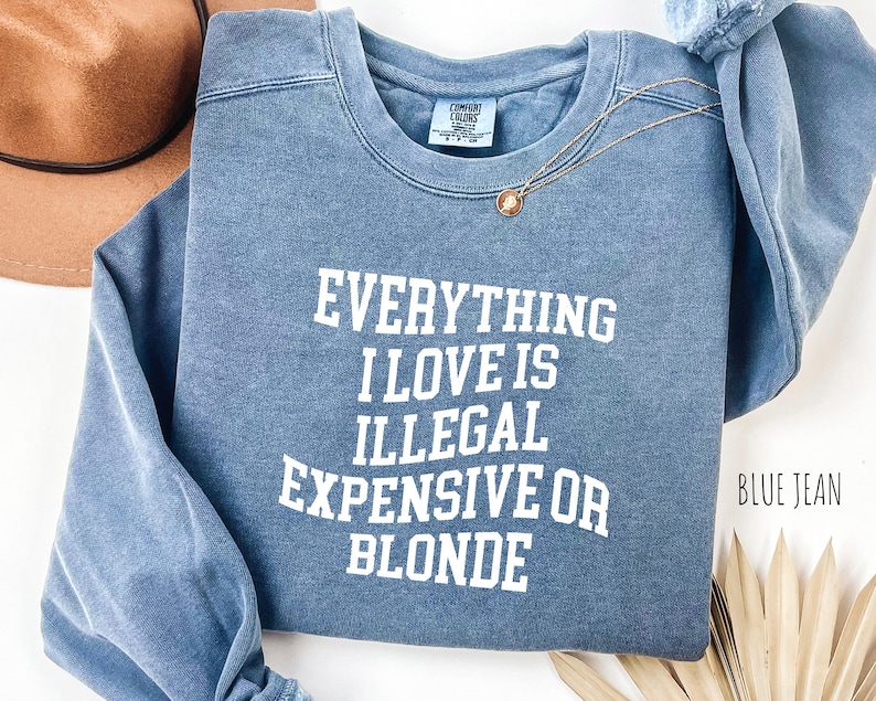 Pu&ograve; includere: Felpa blu jeans con la scritta "EVERYTHING I LOVE IS ILLEGAL EXPENSIVE OR BLONDE" in lettere bianche. Sono visibili anche una collana d'oro e un cappello marrone.