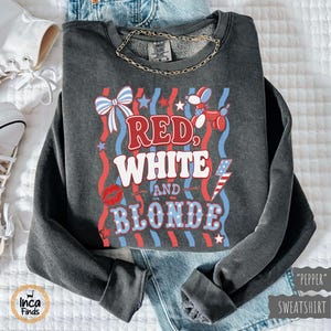 May include: A dark grey sweatshirt featuring the text "Red, White, and Blonde" in red, white, and blue. The design includes a bow, stars, a balloon dog, a lightning bolt, and a pair of red lips. A gold chain necklace is visible.