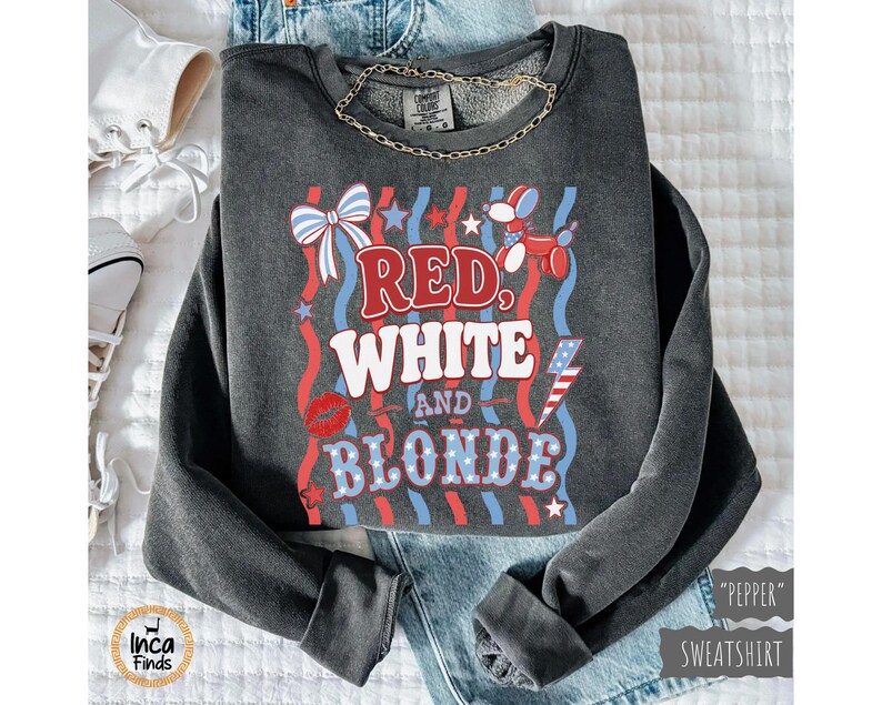 May include: A dark grey sweatshirt featuring the text "Red, White, and Blonde" in red, white, and blue. The design includes a bow, stars, a balloon dog, a lightning bolt, and a pair of red lips. A gold chain necklace is visible.