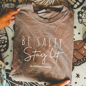 以下が含まれることがあります： エスプレッソ色のTシャツ。白い筆記体で「BE SALTY Stay Lit」と書かれています。テキスト「MATTHEW 5:13-14」が下にあります。シャツは折りたたまれ、手で持たれています。