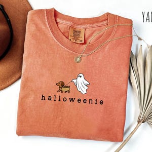 May include: A yam-colored t-shirt with a crew neck and the text "halloweienie". The shirt features a cartoon dachshund dog on a leash with a ghost. A gold necklace is draped across the shirt.