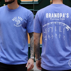 May include: A periwinkle blue short-sleeve t-shirt. The front features a white graphic of a fish with the text "REEL COOL Grandpa". The back displays a white graphic of a fishing rod with lines hanging down, each ending in a lure and a name.