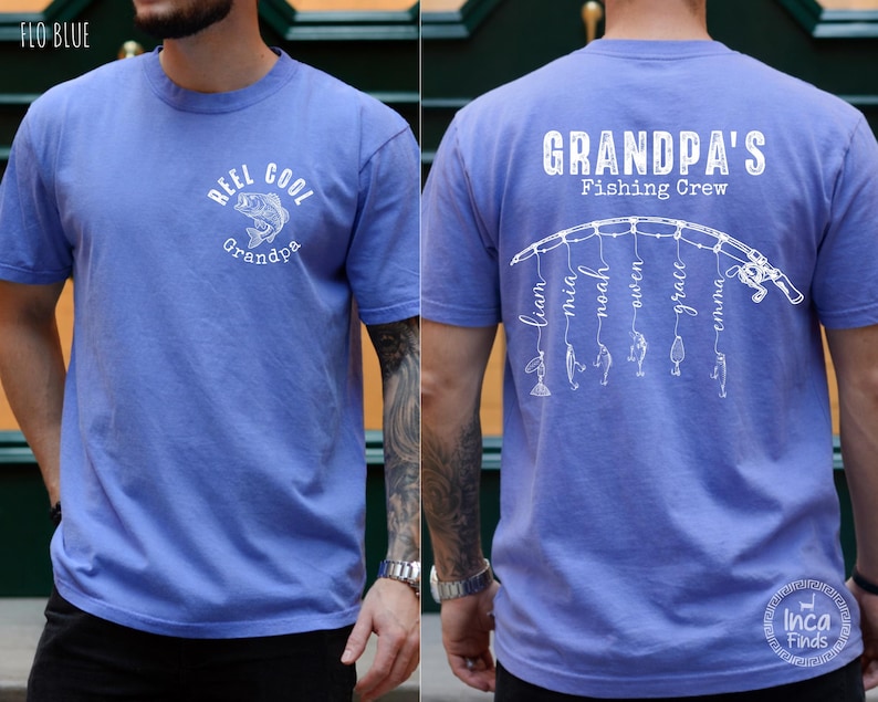 May include: A periwinkle blue short-sleeve t-shirt. The front features a white graphic of a fish with the text "REEL COOL Grandpa". The back displays a white graphic of a fishing rod with lines hanging down, each ending in a lure and a name.