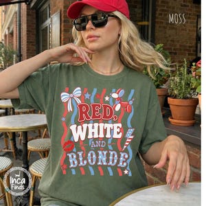 May include: Olive green t-shirt with the text "RED, WHITE, AND BLONDE" in red, white, and blue. The shirt features a bow, a balloon dog, a kiss, and an American flag graphic. The word "MOSS" is in the upper right corner.