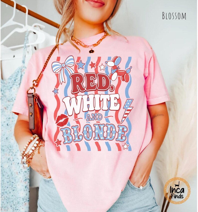 May include: Pale pink t-shirt featuring the text "RED, WHITE, and BLONDE" in red and blue. The design includes a bow, stars, a balloon dog, a kiss, and a lightning bolt. The word "BLOSSOM" is printed in the top right corner.