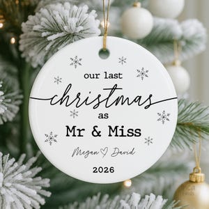 May include: White ceramic Christmas ornament with gold string. The ornament has the text "our last christmas as Mr & Miss" with the names "Megan & David" and the year "2026" in black script. Snowflake designs are scattered around the text.