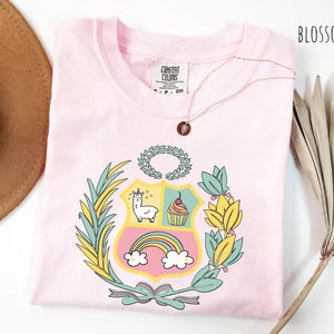 May include: A light pink t-shirt with a crest design featuring a llama, cupcake, and rainbow. The shirt has a crew neck and short sleeves. The word "BLOSSOM" is printed in black on the top right corner.
