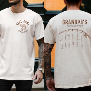 May include: An ivory t-shirt featuring brown graphics. The front displays a fish illustration with the text "REEL COOL Grandpa". The back shows a fishing rod graphic with the text "GRANDPA'S Fishing Crew" and names hanging from the line.