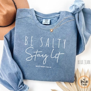 May include: A blue jean-colored sweatshirt with the words "BE SALTY Stay Lit" in white script. The text "MATTHEW 5:13-14" is below the main text. A gold necklace and a brown hat are also visible.